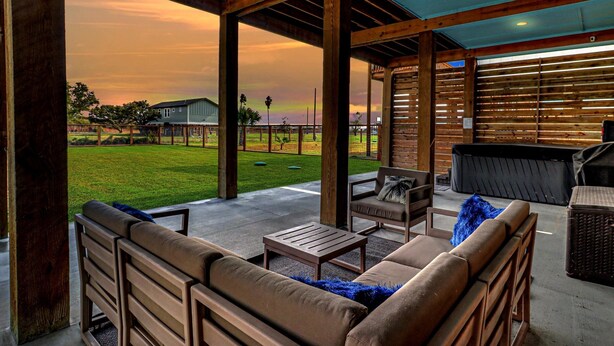 An outdoor patio with wooden furniture, a barbecue grill, and a view of a grassy field and distant buildings. - Nautical Nest 2 Bedroom Home (Rockport)