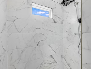 Un baño con baldosas de mármol blanco, una ventana pequeña con vista al cielo y una zona de ducha con cabezal de ducha manual.