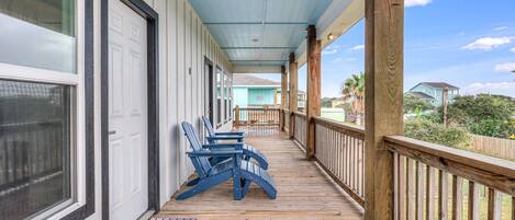 A covered porch with wooden railings, two blue Adirondack chairs, and a view of a residential area with houses and greenery.