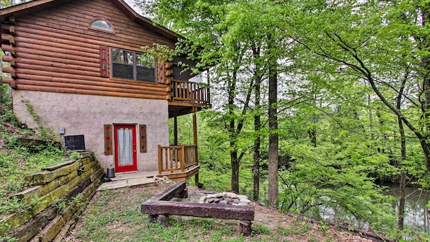 Une cabane en rondins avec une porte rouge et un balcon donnant sur une zone boisée, près d’une étendue d’eau.