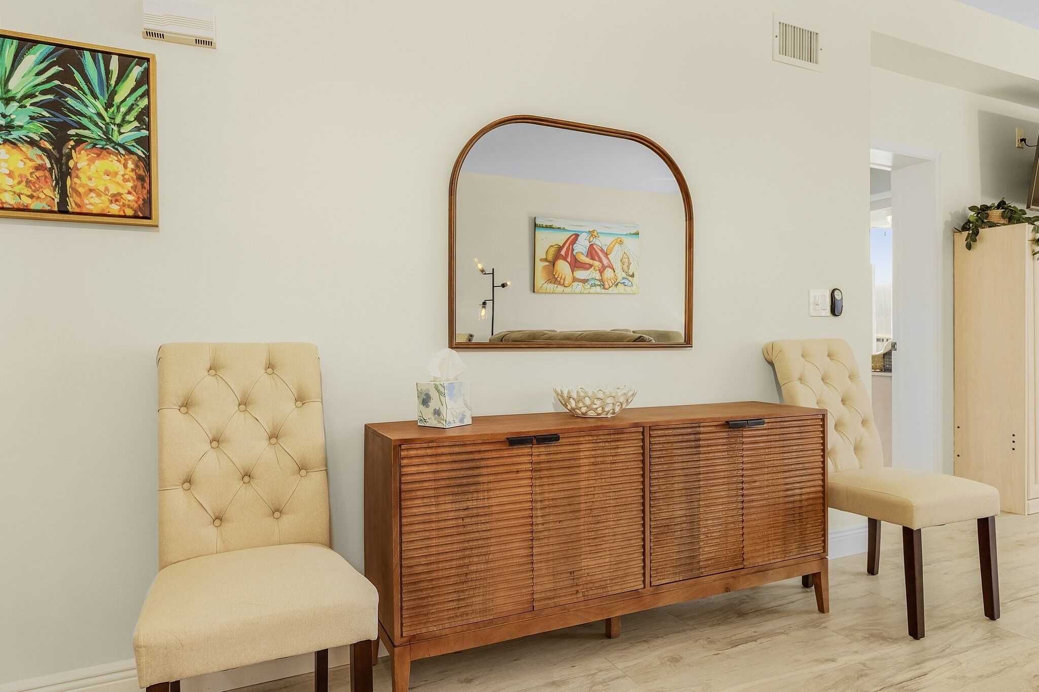 A room with a wooden cabinet, two cushioned chairs, a mirror, and a painting of pineapples.