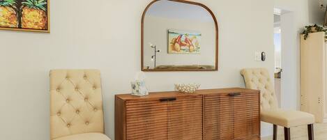 A room with a wooden cabinet, two cushioned chairs, a mirror, and a painting of pineapples.