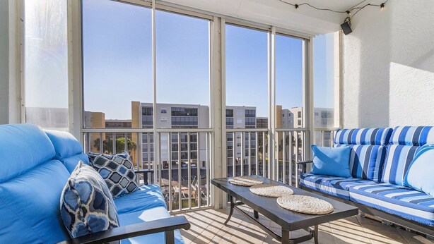 Un balcón con dos sofás azules, una mesita y vistas a edificios a través de amplias ventanas.