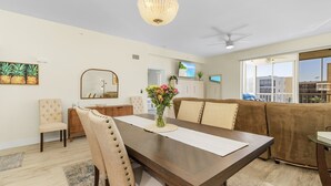 Comedor con una mesa de madera, sillas tapizadas en beige, un florero con flores, una araña y un televisor en la pared.