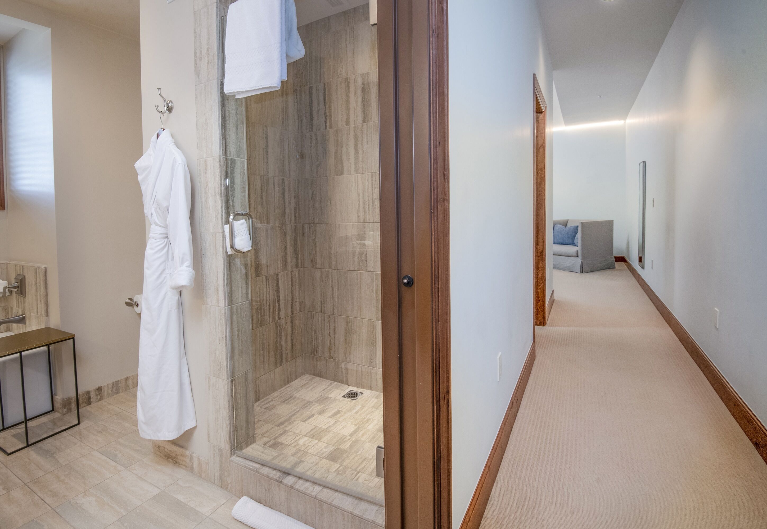 Un baño moderno con ducha envuelta en vidrio, una bata blanca colgada de un perchero y una puerta de madera que da a un pasillo.