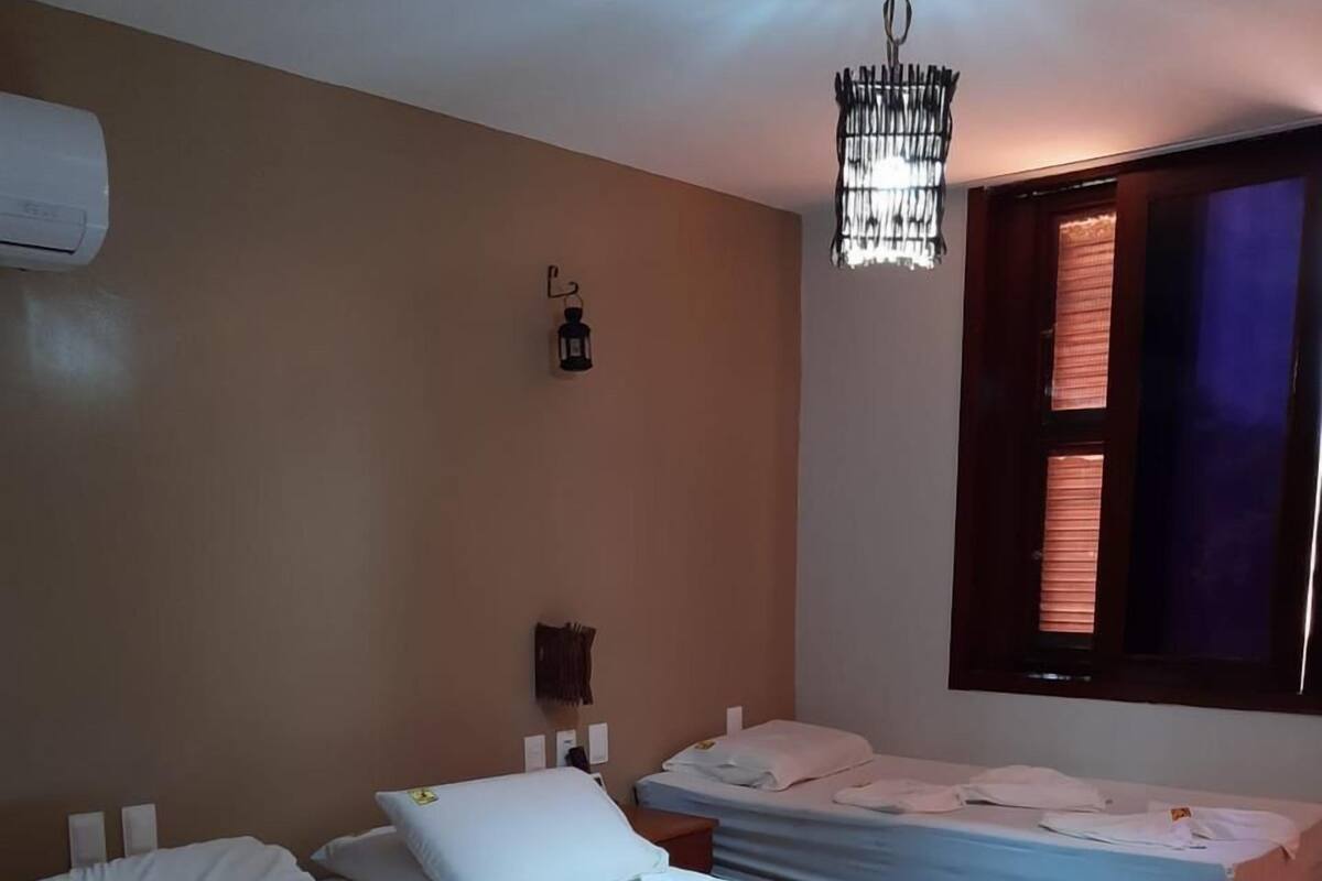 Une chambre d’hôtel avec deux lits, un luminaire suspendu, un climatiseur fixé au mur et une fenêtre avec des stores.