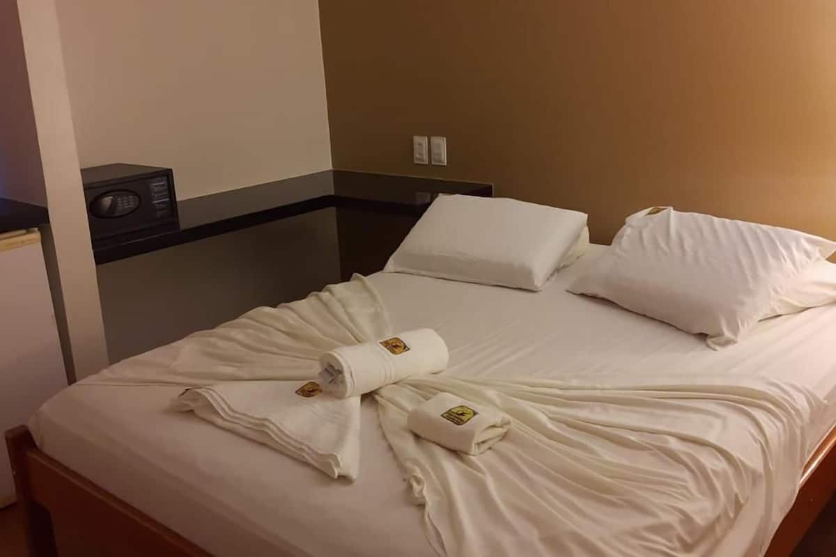 Une chambre d’hôtel avec un lit, du linge de lit blanc, une serviette, une table de chevet, une radio et un climatiseur fixé au mur.