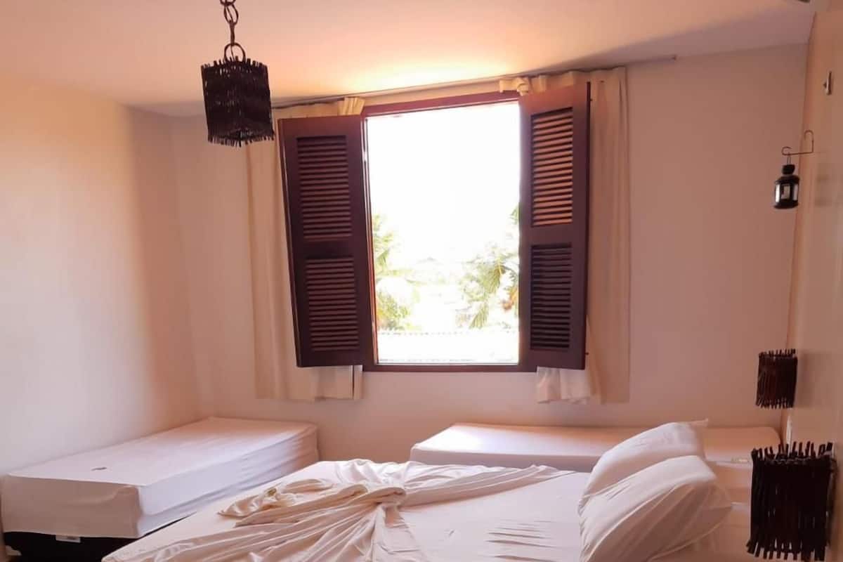 Une chambre avec un lit en bois, une table de chevet, une petite table d’appoint, une fenêtre avec des volets et un luminaire suspendu.