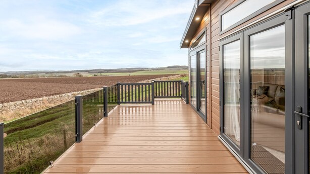 A wooden deck with glass doors leading to a room, overlooking a field. - Lodge No 23 | Hot Tub | St Andrews Lodge Park (Strathkinness)