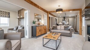 A modern living room with a wooden coffee table, sofas, and a kitchen in the background. - Lodge No 23 | Hot Tub | St Andrews Lodge Park (Strathkinness)