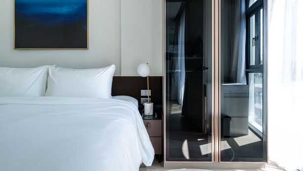Une chambre d’hôtel moderne avec un grand lit, une table de chevet avec une lampe et une vue sur un balcon, accessible par une porte vitrée.