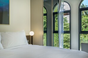 Een hotelkamer met een groot bed, een tafellamp met bolvormige kap en grote ramen met uitzicht op groen buiten.
