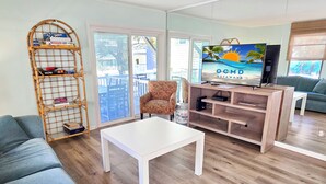 Una sala de estar con un sofá, una mesa de centro, un televisor sobre un soporte de madera, un estante de mimbre con juegos de mesa y vistas a través de una puerta corredera.
