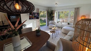 A modern living room with a dining area, a television, and a large window overlooking a garden. - Forest view holiday house with private jacuzzi (Hoenderloo)