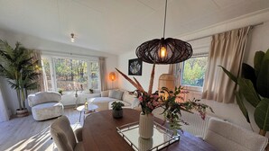A modern living room with a wooden dining table, a glass-top coffee table, and a hanging light fixture. There are potted plants and a sofa in the room. - Forest view holiday house with private jacuzzi (Hoenderloo)