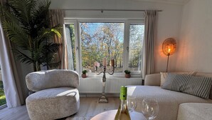 A cozy living room with a sofa, a small table with a wine bottle and glasses, a potted plant, and a view of trees through large windows. - Forest view holiday house with private jacuzzi (Hoenderloo)