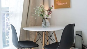 A modern room with a round white table, two black chairs, a vase with flowers, a poster on the wall, and a window with curtains.