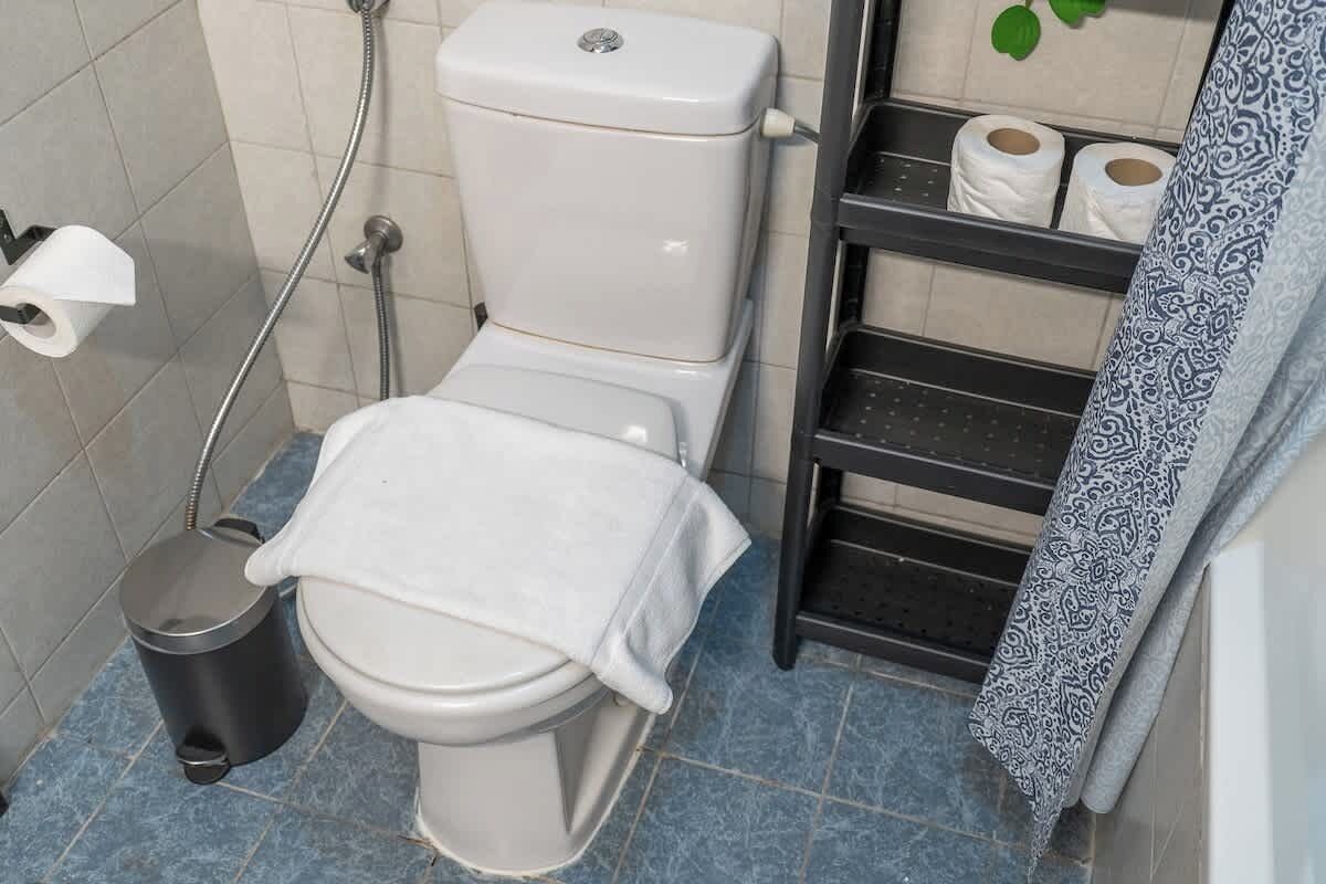 A bathroom with a toilet, a trash bin, a shelf with toilet paper, and a patterned towel.