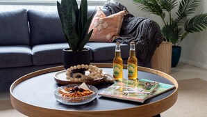 Una mesa redonda de madera con una revista, dos botellas de cerveza, un cuenco de palitos de pretzel, una planta pequeña y un objeto decorativo.