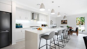 Una cocina moderna con armarios blancos, un refrigerador negro, una isla central con taburetes y luminarias colgantes, además de un comedor con vistas a plantas tropicales.
