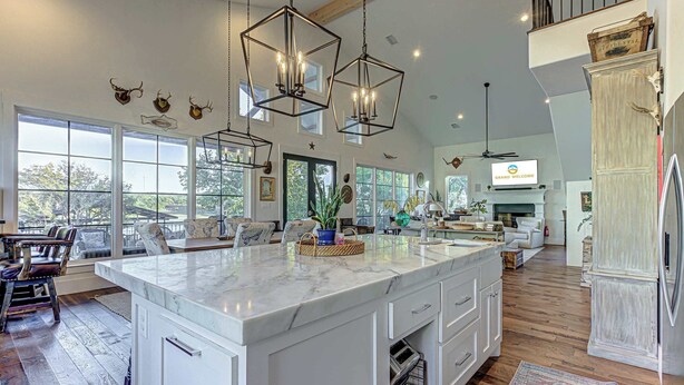 A spacious kitchen with a large island, modern pendant lights, and a view of the outdoors through large windows. - LBJ Waterfront Escape | Private Dock & sleeps 16! (Kingsland)