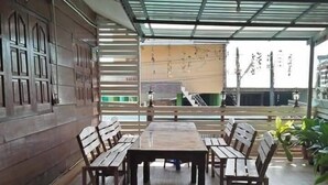 An outdoor patio with a wooden table and benches, a metal roof, and a view of buildings in the background. - Napakarn (Mae Chan)