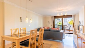 Een moderne woonkamer met een glazen eettafel, houten stoelen, een grijze bank en een balkon met tuinmeubilair.