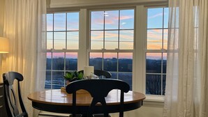 A dining area with a round table and chairs, a potted plant, and a view of a sunset through large windows. - Sunset Serenity Suite (Myerstown)