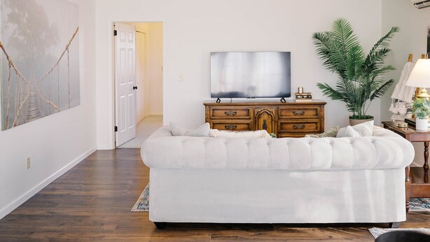 A living room with a white sofa, a wooden cabinet with a flat-screen TV, a large plant, and a door leading to another room. - Sunset Serenity Suite (Myerstown)