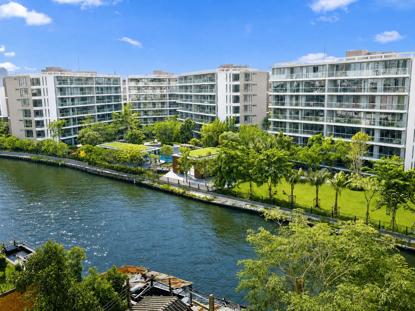 Ein moderner Wohnkomplex mit mehreren Stockwerken, einem Fluss im Vordergrund und einer gepflegten Grünanlage mit Bäumen.