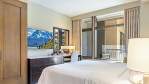 A hotel room with a large bed, a desk with a chair, a dresser, a large window with curtains, and a mountain view painting on the wall.
