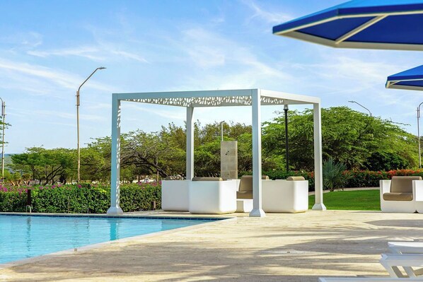 Un espace aménagé au bord de la piscine, avec une pergola blanche, des chaises longues et un ciel d’un bleu limpide.