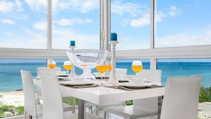 Une table à manger pour quatre personnes, avec des chaises blanches, placée près d’une grande fenêtre donnant sur l’océan.