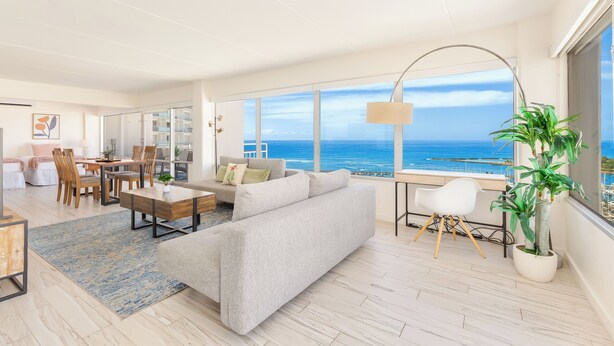 A spacious living area with a large window overlooking the ocean, a sofa, a dining table, and a potted plant.