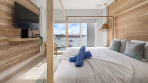 A modern bedroom with a large bed, a TV mounted on a wooden wall, and a view of the water outside. - Beach Vibes Ocean View Condo (Oranjestad)