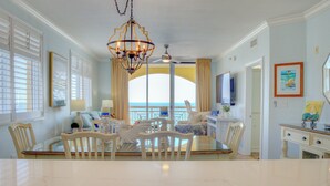 A dining area with a marble table, white chairs, a chandelier, and a view of the ocean through a large window. - Azure 601 | Gulf-Front 4BR Corner Unit | FREE Beach Service! (Fort Walton Beach)