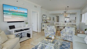 A spacious living room with a large flat-screen TV, comfortable seating, and a modern kitchen in the background. - Azure 601 | Gulf-Front 4BR Corner Unit | FREE Beach Service! (Fort Walton Beach)