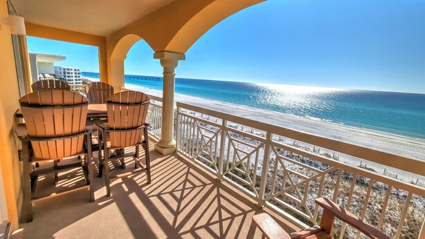 A balcony with a view of the beach and ocean, featuring wooden chairs and a table. - Azure 601 | Gulf-Front 4BR Corner Unit | FREE Beach Service! (Fort Walton Beach)