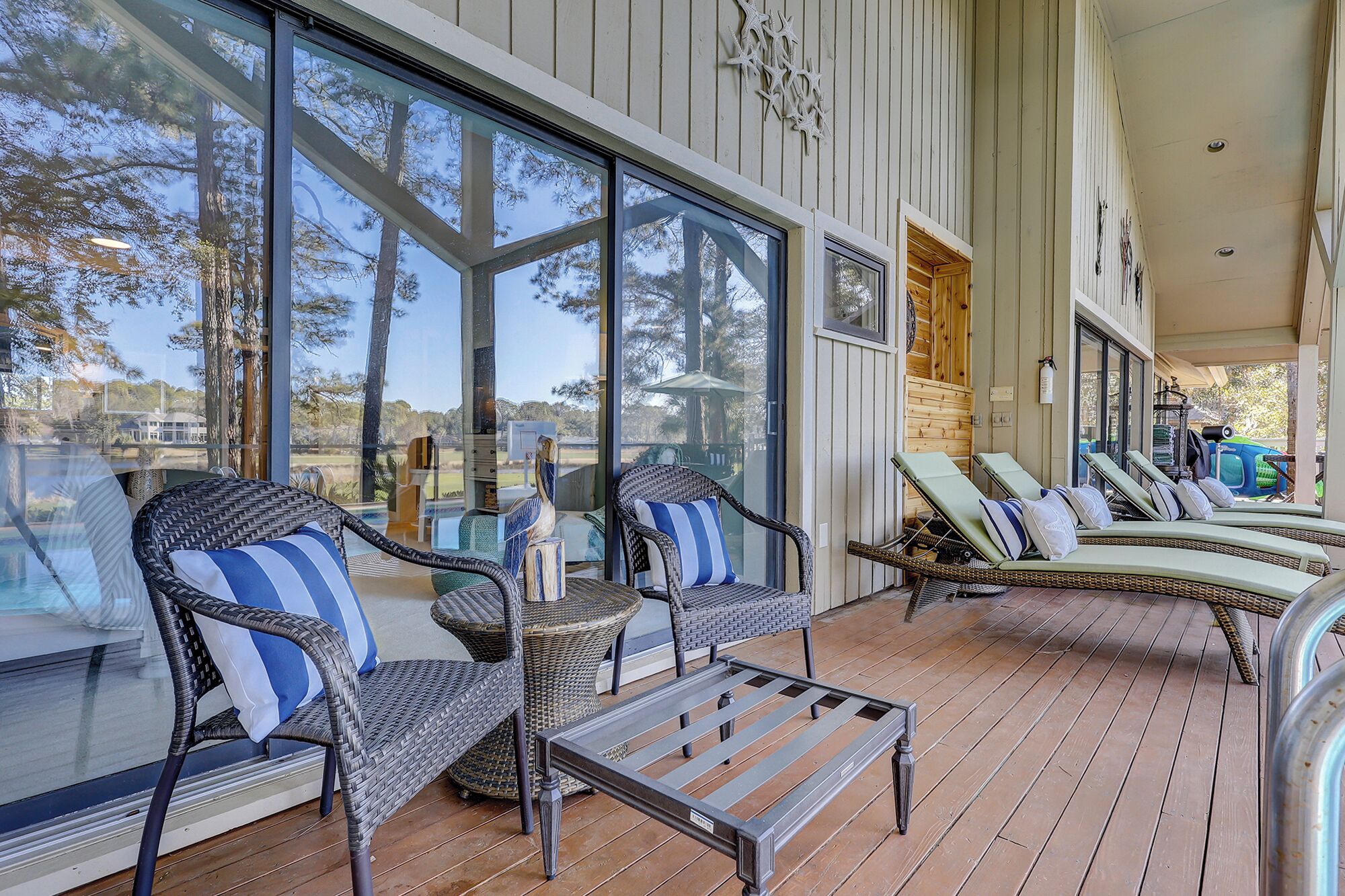 A covered outdoor seating area with wicker chairs and lounge chairs, large windows, and a wooden deck.