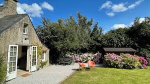 A stone house with a gravel driveway, a wooden door, and a small garden with pink flowers and a table with chairs. - Charming cottage, sea and garden, close to the D-Day landing beaches, sleeps 6 (Quinéville)