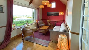 A cozy room with a wooden ceiling, a sofa, a table, and chairs. There is a window with curtains and a view of greenery outside. - Charming cottage, sea and garden, close to the D-Day landing beaches, sleeps 6 (Quinéville)