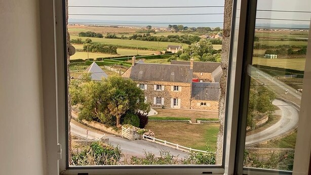 A view from a window overlooking a rural landscape with a stone building, a tree, and a winding road. - Charming cottage, sea and garden, close to the D-Day landing beaches, sleeps 6 (Quinéville)
