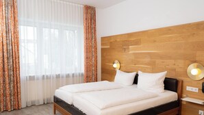 Moderni hotellihuone, jossa on puinen pääty, suuri sänky valkoisilla vuodevaatteilla, yöpöytä lampun kanssa ja ikkuna, jossa on läpikuultavat verhot.