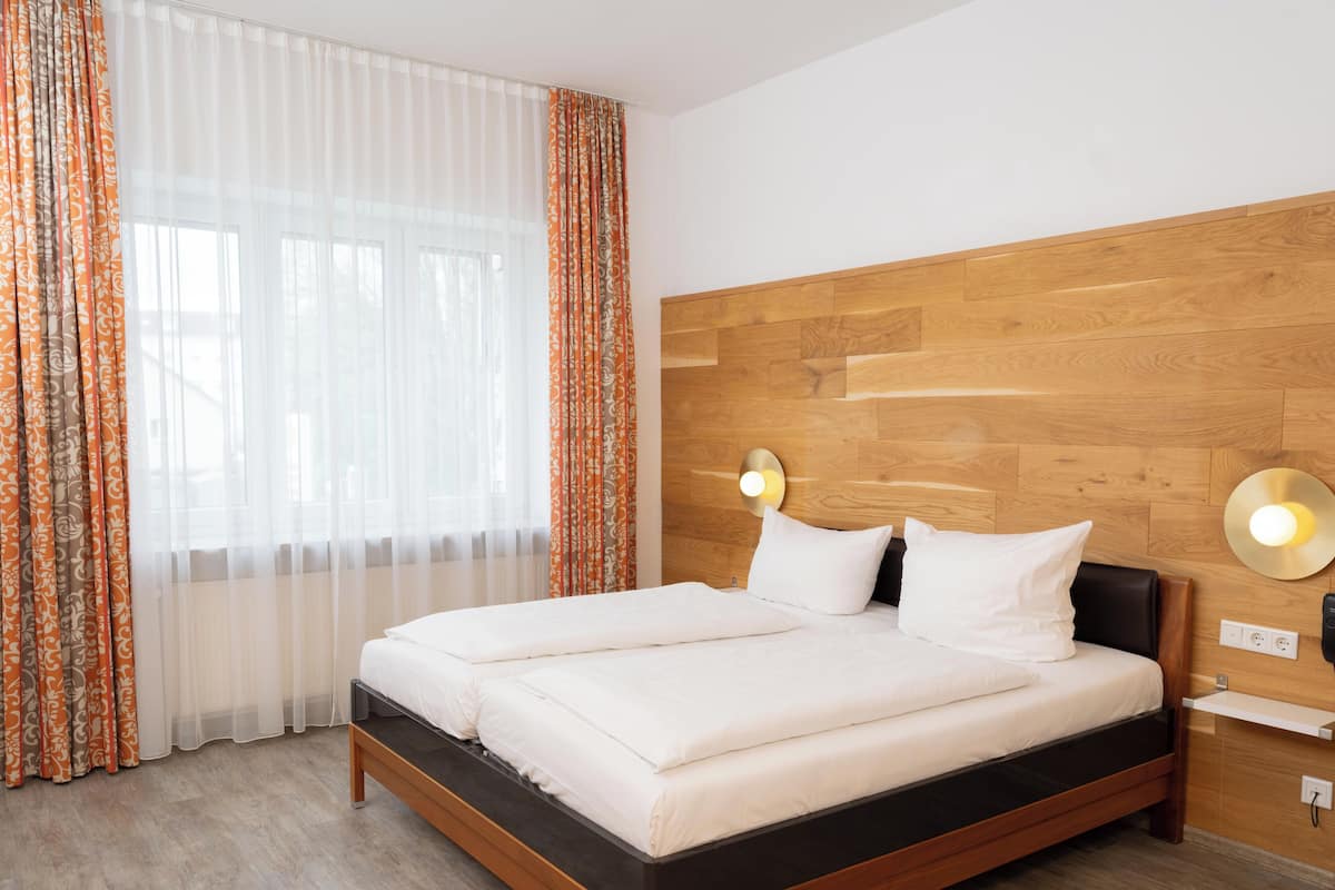 A modern hotel room with a wooden headboard, a large bed with white bedding, a bedside table with a lamp, and a window with sheer curtains.