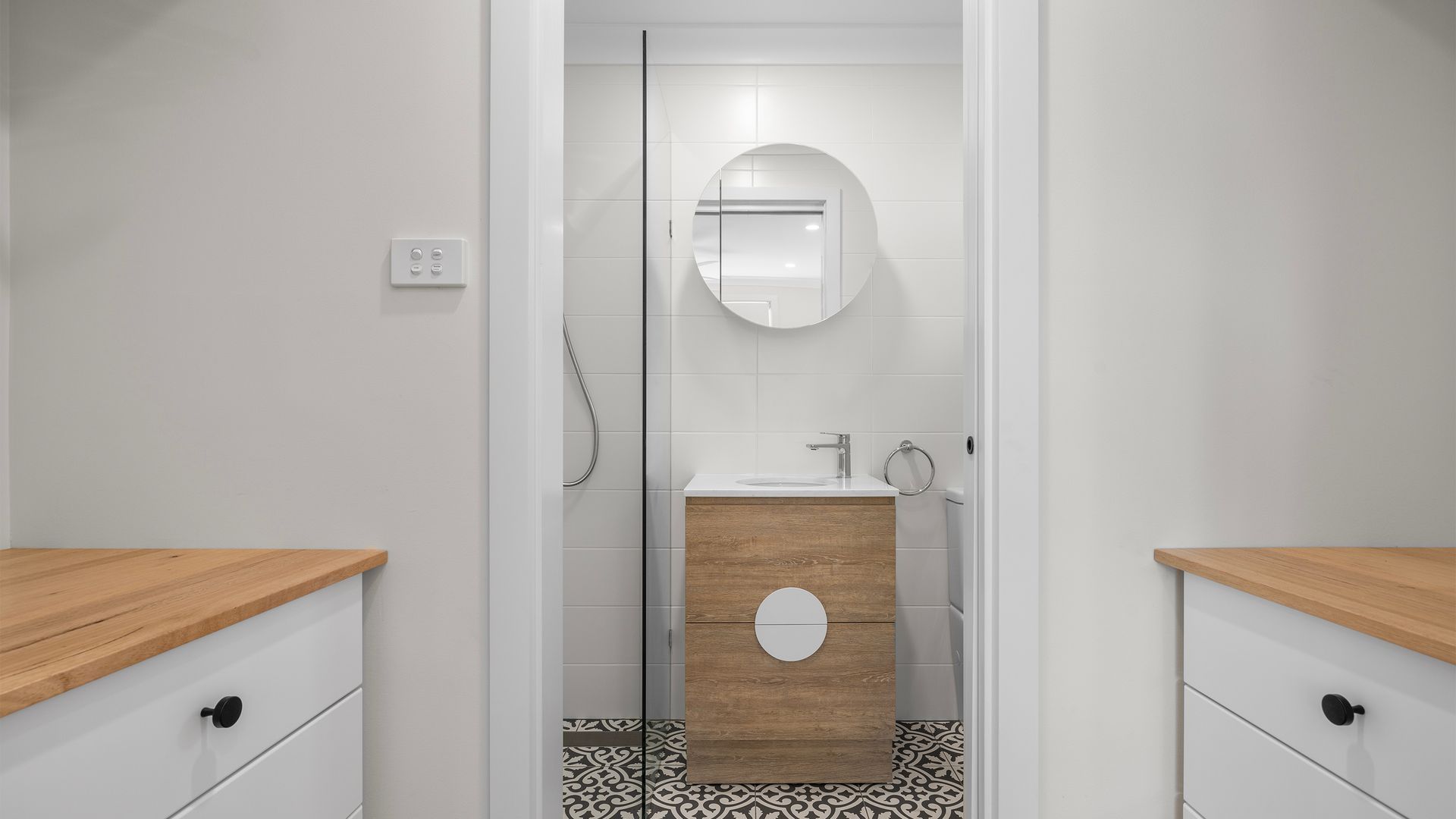 A modern bathroom with a wooden vanity, a round mirror, and a shower area.