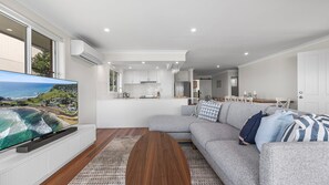 A modern living room with a large flat-screen TV, a grey sofa with patterned cushions, a wooden coffee table, and a view of the kitchen and dining area.