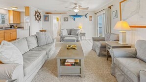 A living room with a sofa, a coffee table with books, a ceiling fan, and a view of the kitchen and outside. - Soundfront Home in Frisco with Hot Tub, Private Dock and Windsurfing Access (Frisco)