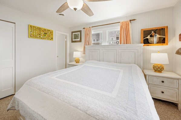 Une chambre à coucher avec un grand lit, deux tables de chevet avec des lampes, un ventilateur de plafond, une fenêtre avec des rideaux et une œuvre d’art murale.