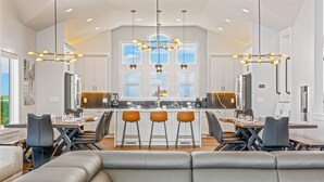 A modern kitchen with a central island, bar stools, and a dining area with a sofa. - Premier Soundfront w/ Pool, Hot Tub, Elevator, EV Charger, Theater & Game Rooms (Avon)