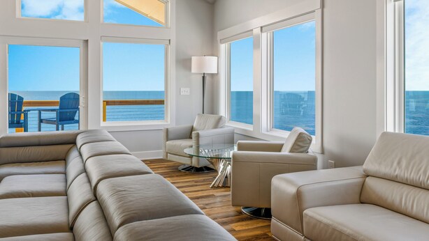 A spacious living room with large windows offering ocean views, a comfortable sofa, a glass coffee table, and a floor lamp. - Premier Soundfront w/ Pool, Hot Tub, Elevator, EV Charger, Theater & Game Rooms (Avon)